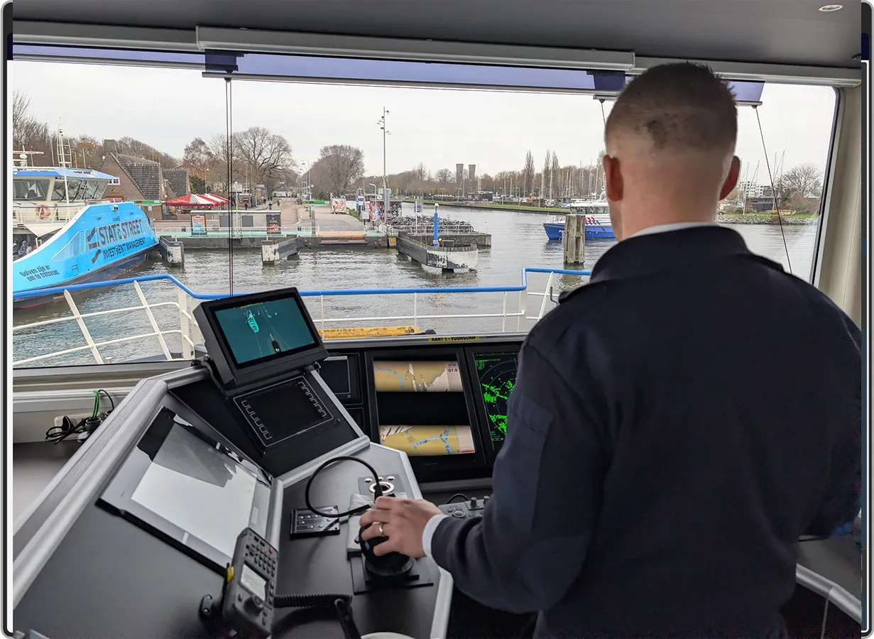 Een persoon bestuurt een veerboot met behulp van navigatiebediening en schermen, terwijl hij een aanlegsteiger nadert met boten die langs een met bomen omzoomde waterkant liggen.