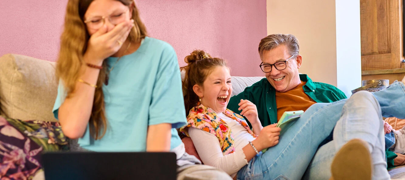 A man, young girl, and a woman laughing together on a couch, with the woman covering her mouth with her hand.