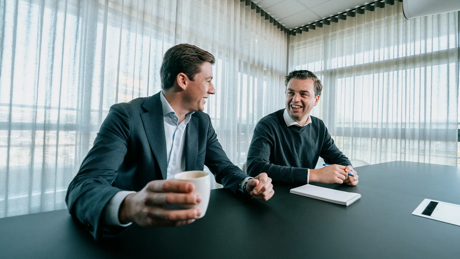 Twee mannen in zakenkleding zitten aan een tafel, pratend en glimlachend. De één houdt een koffiekopje vast en de ander heeft een notitieboekje en een pen.