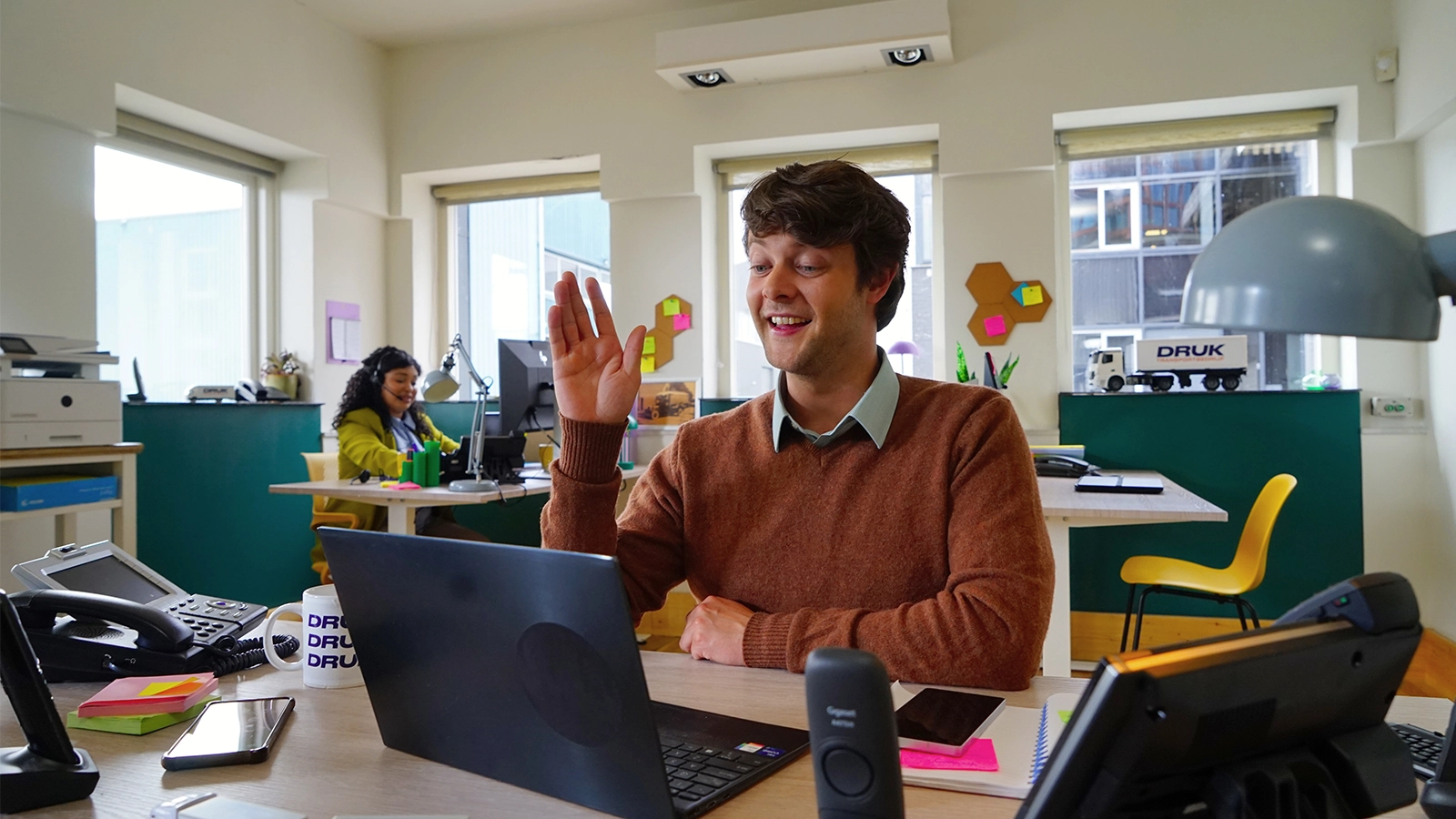 Man in brown sweater waving at laptop during video call in bright office; woman working in background.