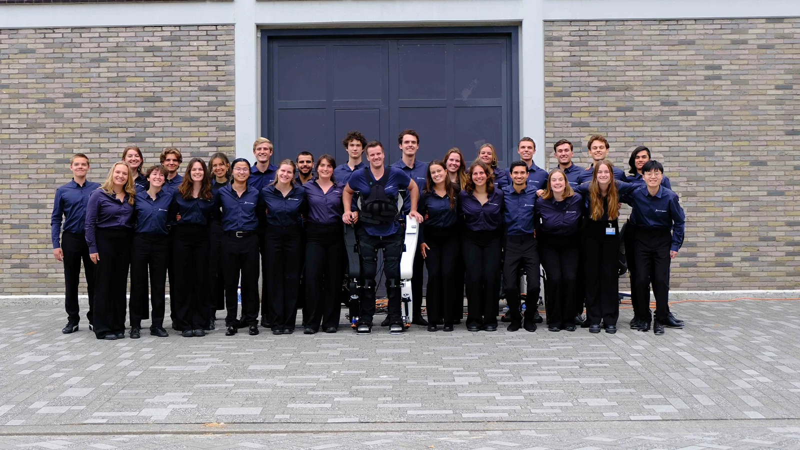 Group of 24 people in matching navy shirts, smiling and standing together in front of a brick wall. One person in the center is wearing an exoskeleton.