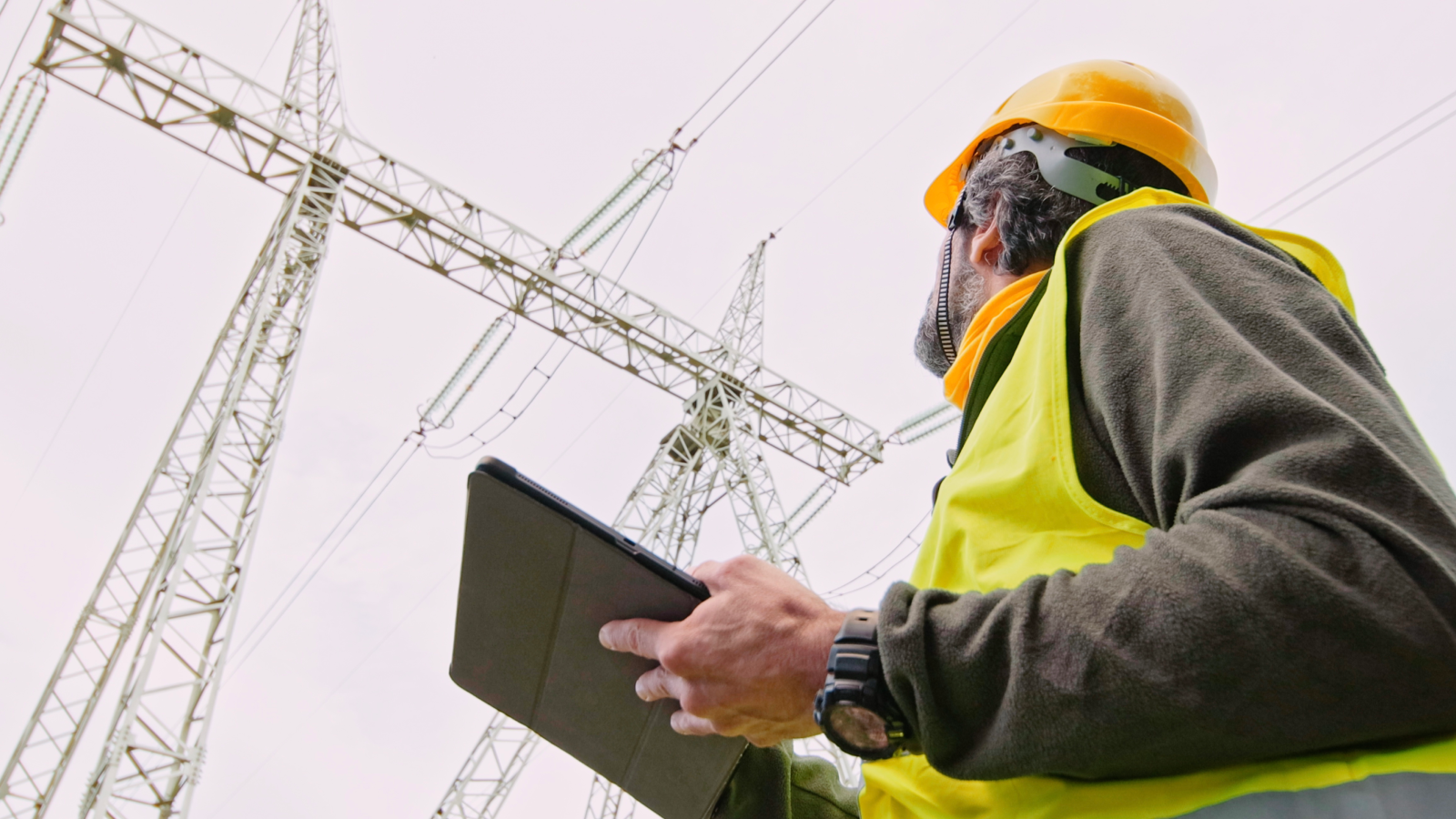 Een werknemer met een geel hesje en een veiligheidshelm houdt een tablet vast en inspecteert elektriciteitsmasten onder een bewolkte hemel. Een werknemer met een geel hesje en een veiligheidshelm houdt een tablet vast en inspecteert elektriciteitsmasten onder een bewolkte hemel.