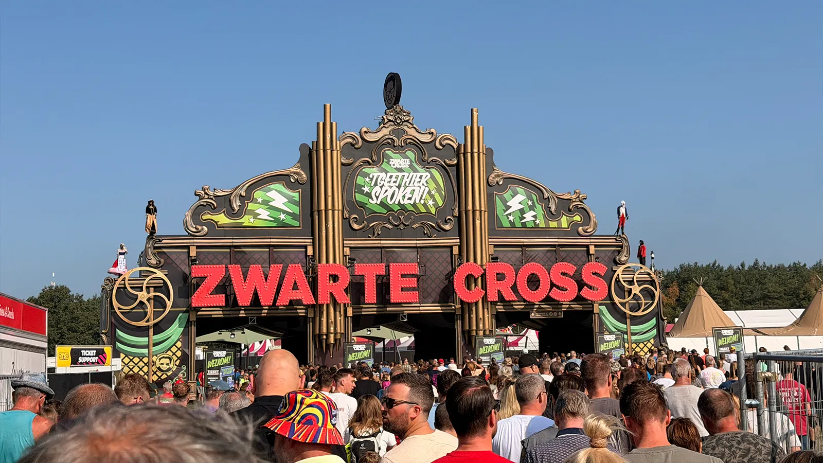 Een menigte verzamelt zich bij de ingang van het Zwarte Cross-festival onder een grote decoratieve boog, waarop de naam “Zwarte Cross” prominent in rode letters staat weergegeven.