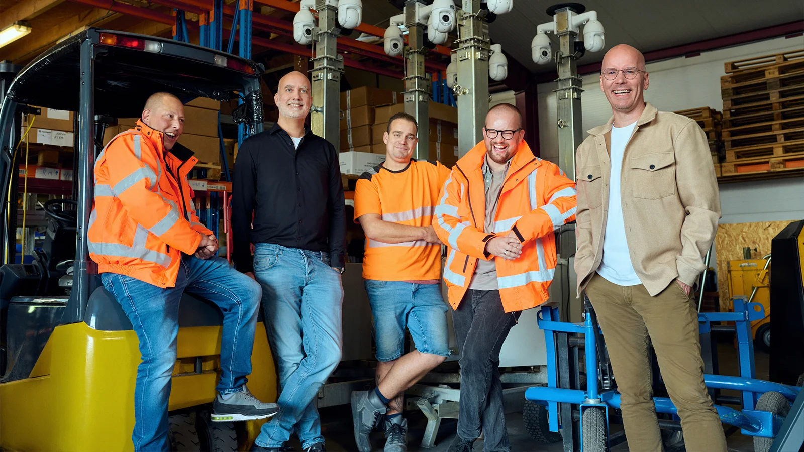 Vijf mannen staan glimlachend in een magazijn, waarvan sommigen feloranje veiligheidsjassen dragen. Ze worden omringd door apparatuur en machines.
