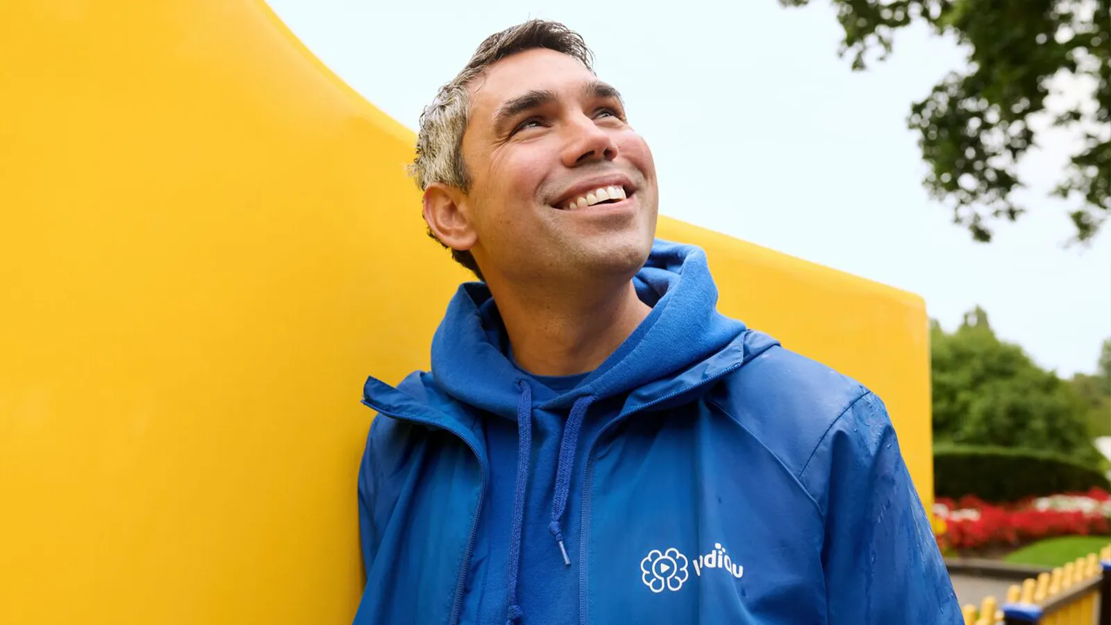 Man in a blue raincoat Man in een blauwe regenjas en hoodie glimlacht terwijl hij omhoog kijkt naar de lucht, staand naast een grote gele constructie, met bomen en een tuin op de achtergrond.hoodie smiling, looking up at the sky, standing near a large yellow structure, with trees and a garden in the background.