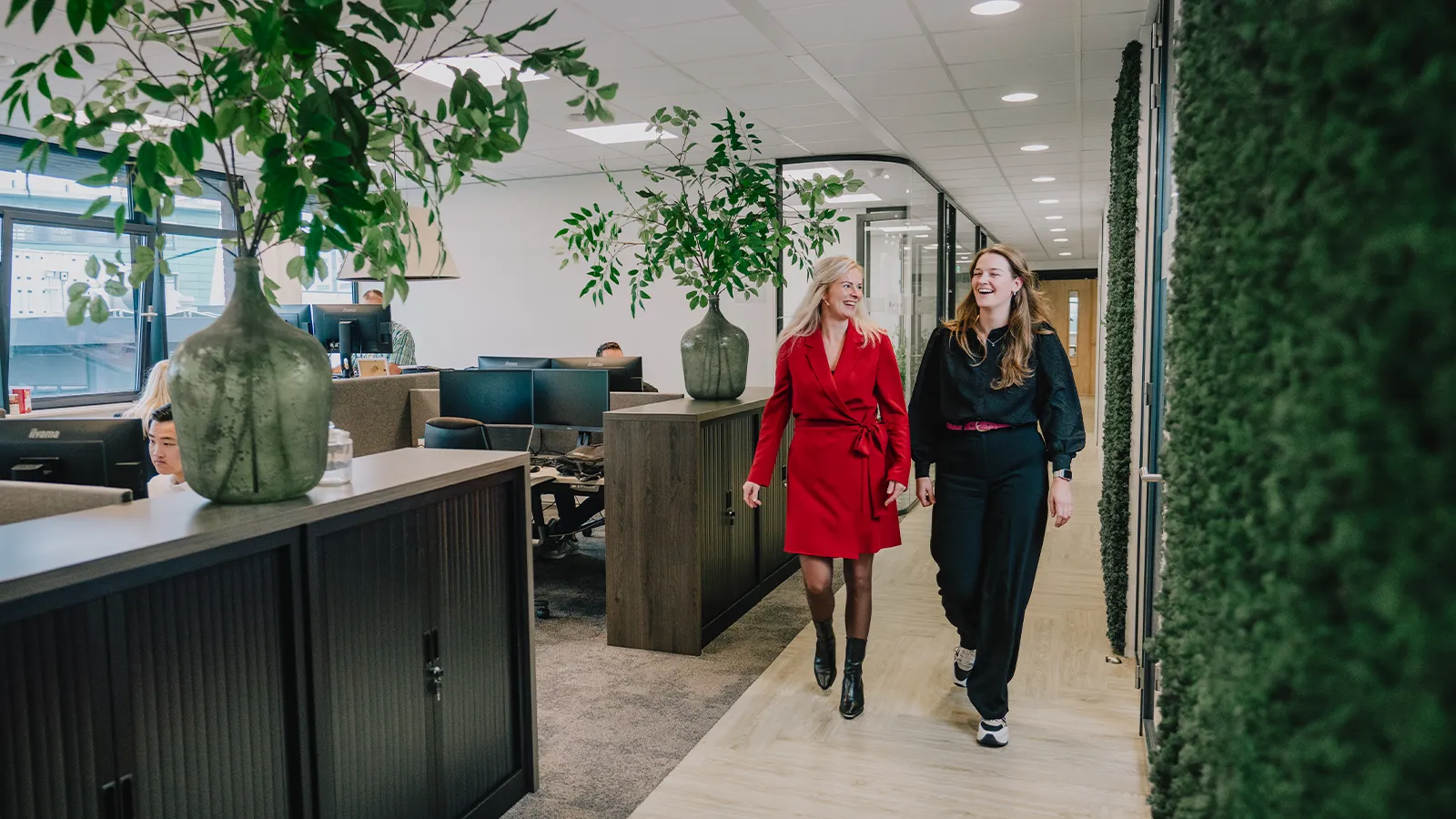 Twee vrouwen lopen en praten door een kantoorgang met modern interieur, grote planten en werkplekken. De één draagt een rode jurk, de ander een zwarte outfit.