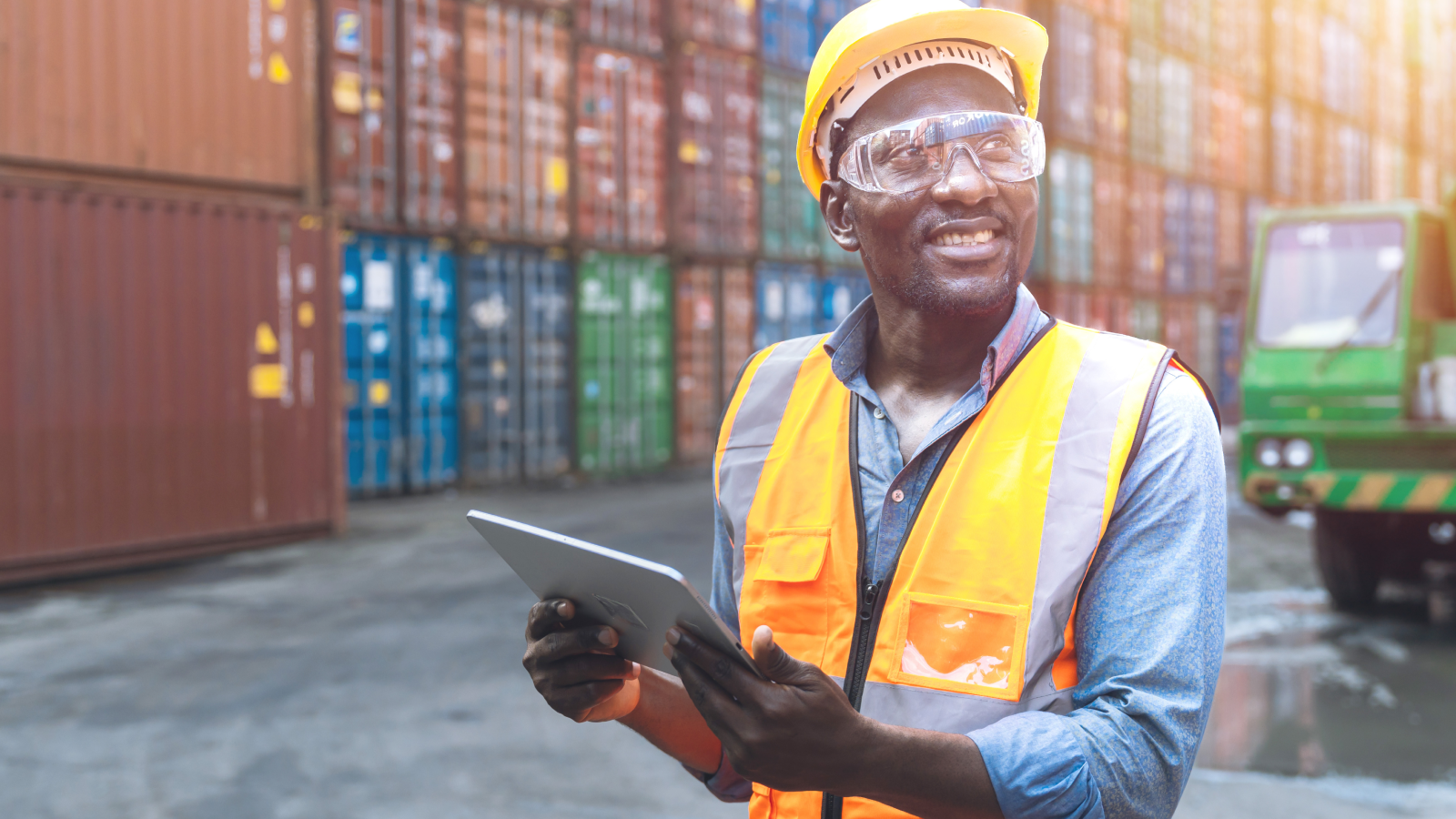 Een werknemer met een veiligheidshelm en veiligheidshesje houdt een tablet vast en staat voor gestapelde zeecontainers op een industriële locatie. Een werknemer met een veiligheidshelm en veiligheidshesje houdt een tablet vast en staat voor gestapelde zeecontainers op een industriële locatie.