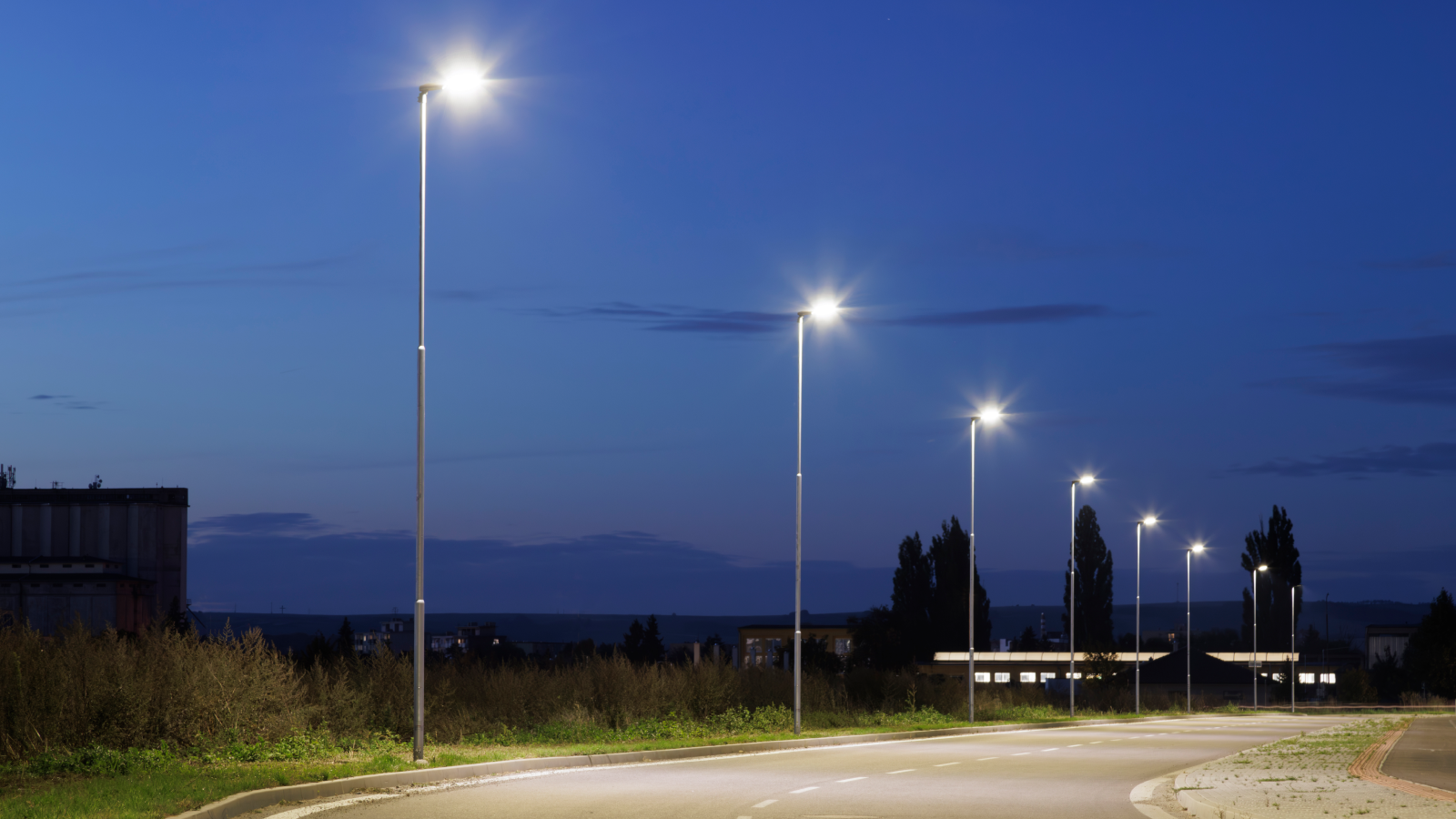 Lantaarnpalen verlichten een bochtige weg tijdens de schemering, met een heldere blauwe lucht en silhouetten van bomen en gebouwen op de achtergrond. Lantaarnpalen verlichten een bochtige weg tijdens de schemering, met een heldere blauwe lucht en silhouetten van bomen en gebouwen op de achtergrond.