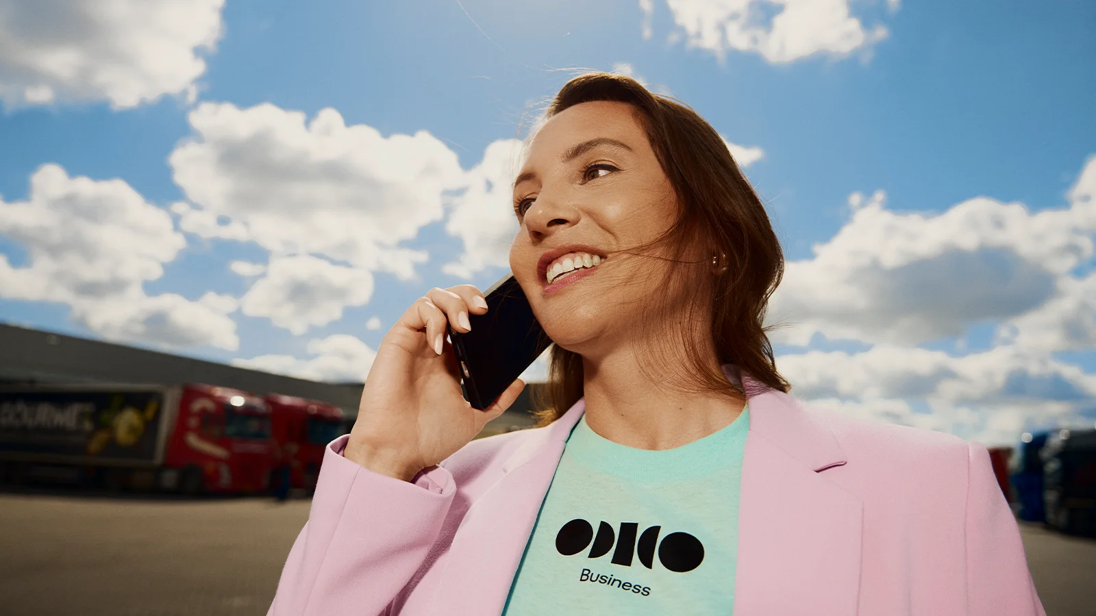 Vrouw in een pastelroze blazer praat glimlachend aan de telefoon onder een gedeeltelijk bewolkte lucht, met vrachtwagens op de achtergrond.
