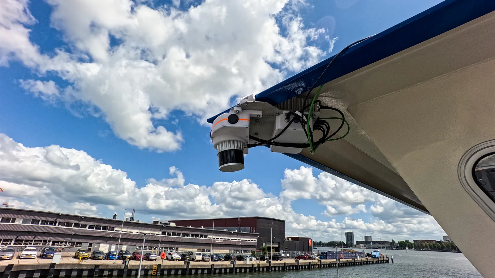 Beveiligingscamera gemonteerd aan de zijkant van een boot die uitkijkt over een aanlegsteiger, met auto’s en een gebouw op de achtergrond onder een halfbewolkte lucht.