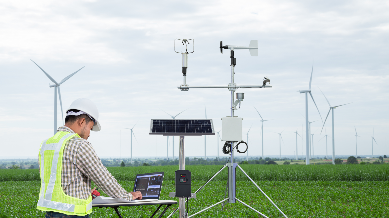 Een ingenieur met een veiligheidshelm en hesje gebruikt een laptop in de buurt van windturbines, met een door zonne-energie aangeduurd weerstation in een veld.