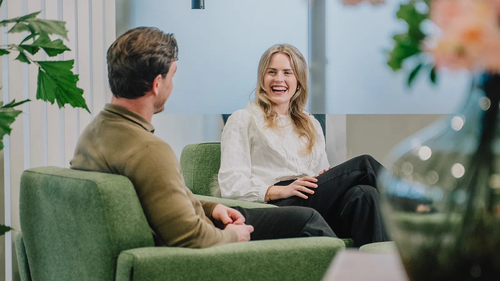 Twee mensen zitten op groene banken en voeren een levendig gesprek, met planten en wazige bloemen op de voorgrond.