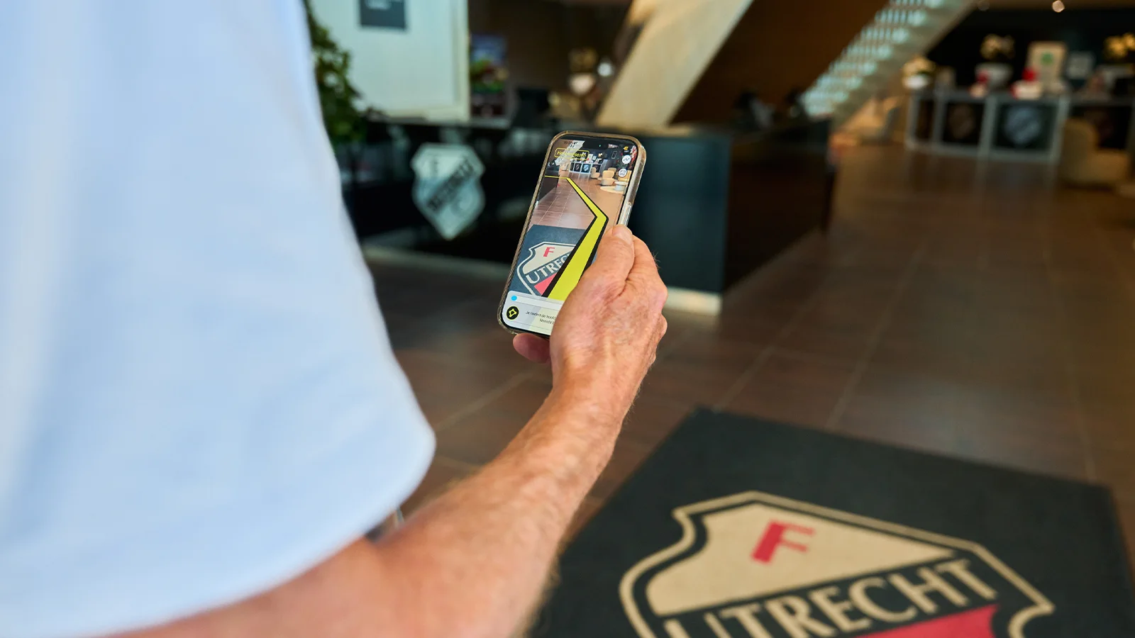 Een persoon houdt een smartphone vast waarop een augmented reality-app wordt weergegeven. De app projecteert een route-aanwijzing over een logo met de tekst “UTRECHT” in een modern interieur.