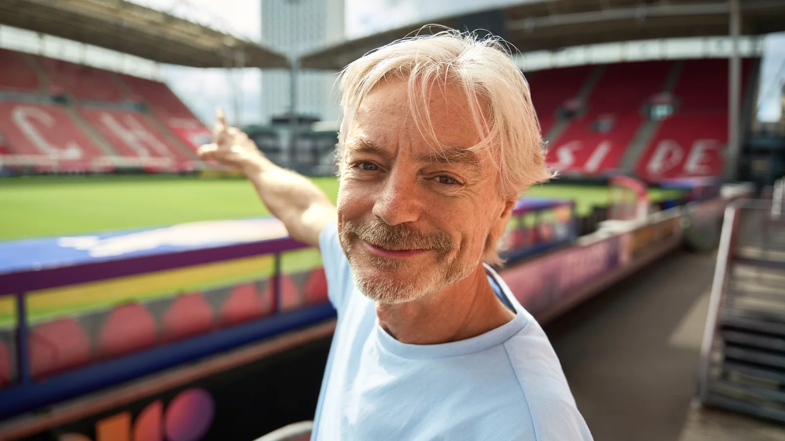 Glimlachende man met grijs haar en een baard wijst naar het veld van een stadion, terwijl hij bij de tribunes van een buitensportarena staat.