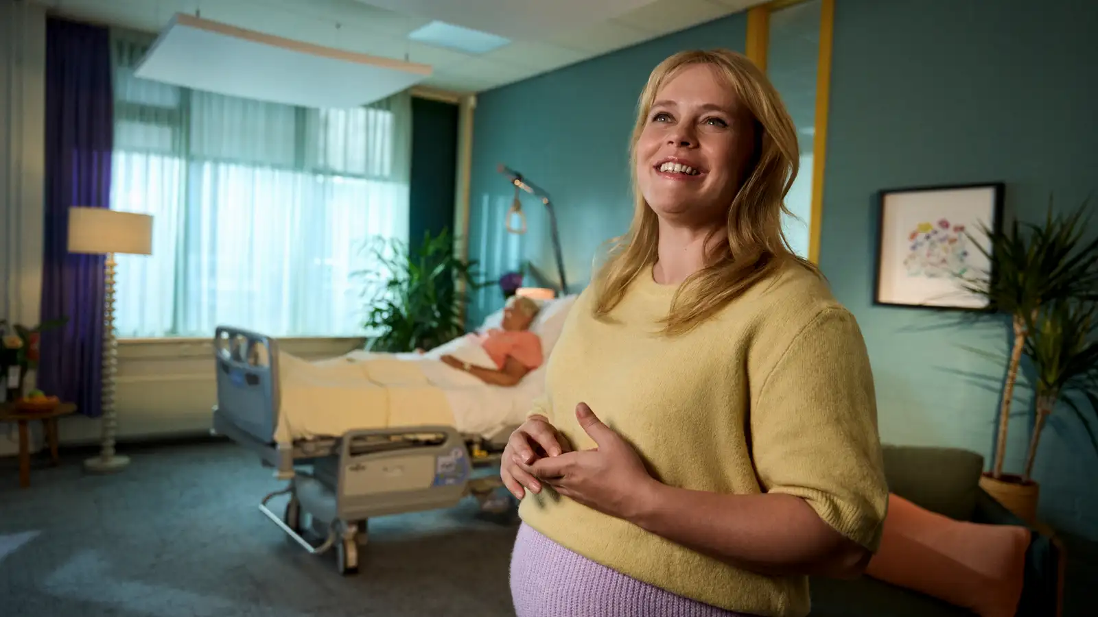 Een glimlachende vrouw staat in een kamer met een ziekenhuisbed en een patiënt op de achtergrond, omringd door planten en zachte verlichting.