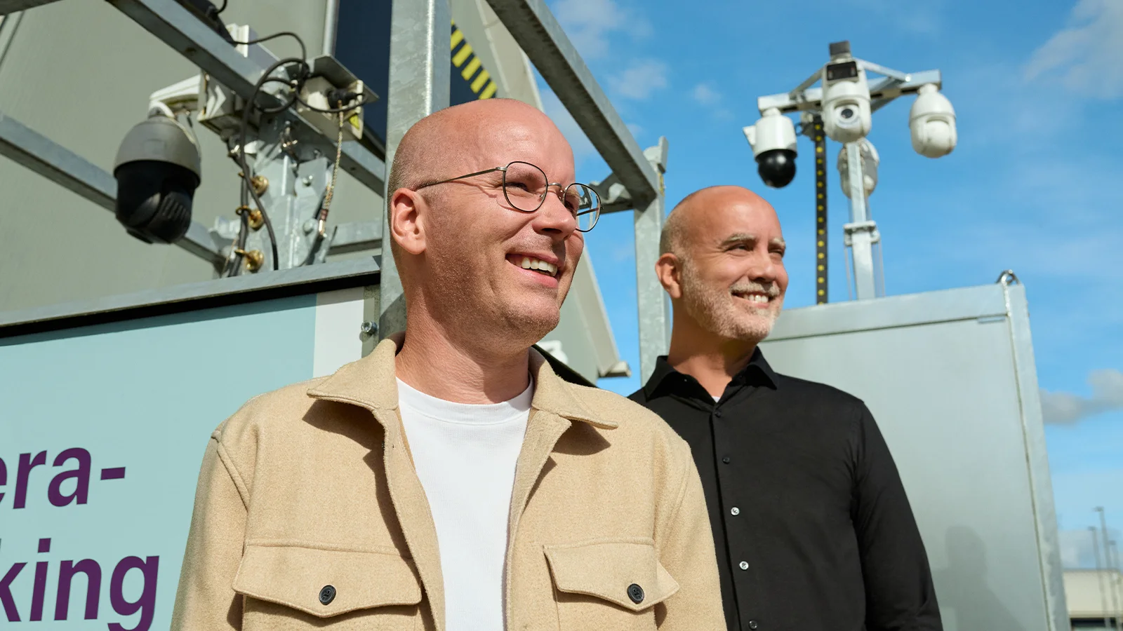 Twee glimlachende mannen, de één in een beige jas en de ander in het zwart gekleed, staan buiten bij bewakingscamera’s en apparatuur onder een blauwe hemel.