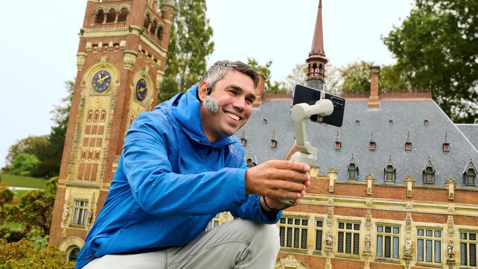 Man in een blauwe jas glimlacht terwijl hij een telefoonstabilisator vasthoudt voor een modelgebouw dat lijkt op een historische klokkentoren met sierlijke architectuur.