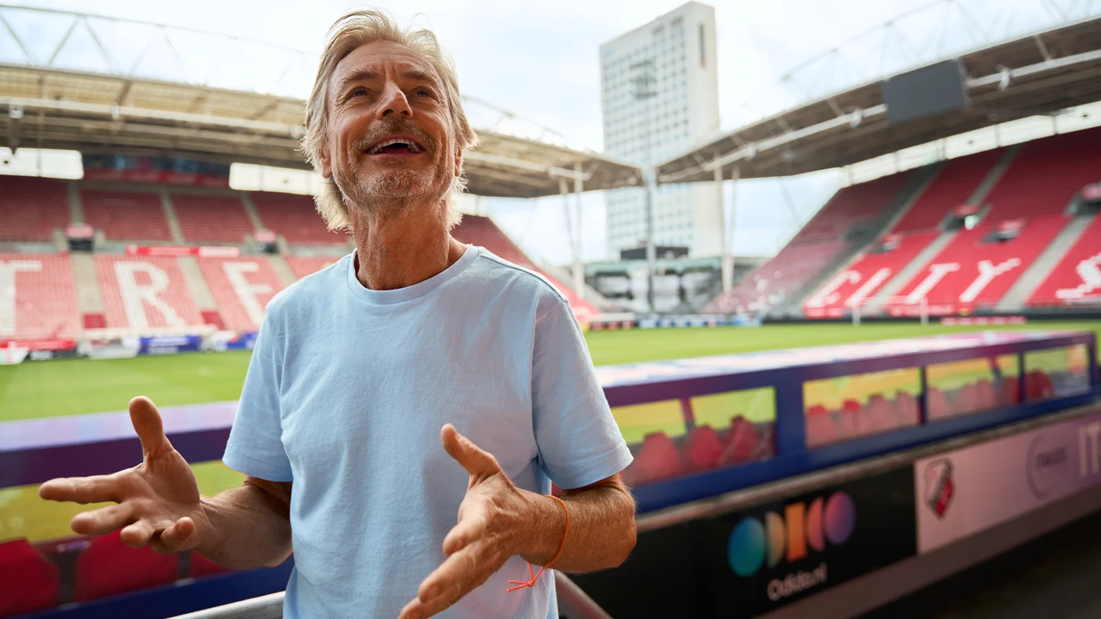 Een man in een lichtblauw overhemd staat gebarend in een stadion met rode tribunezitplaatsen en een modern gebouw op de achtergrond.