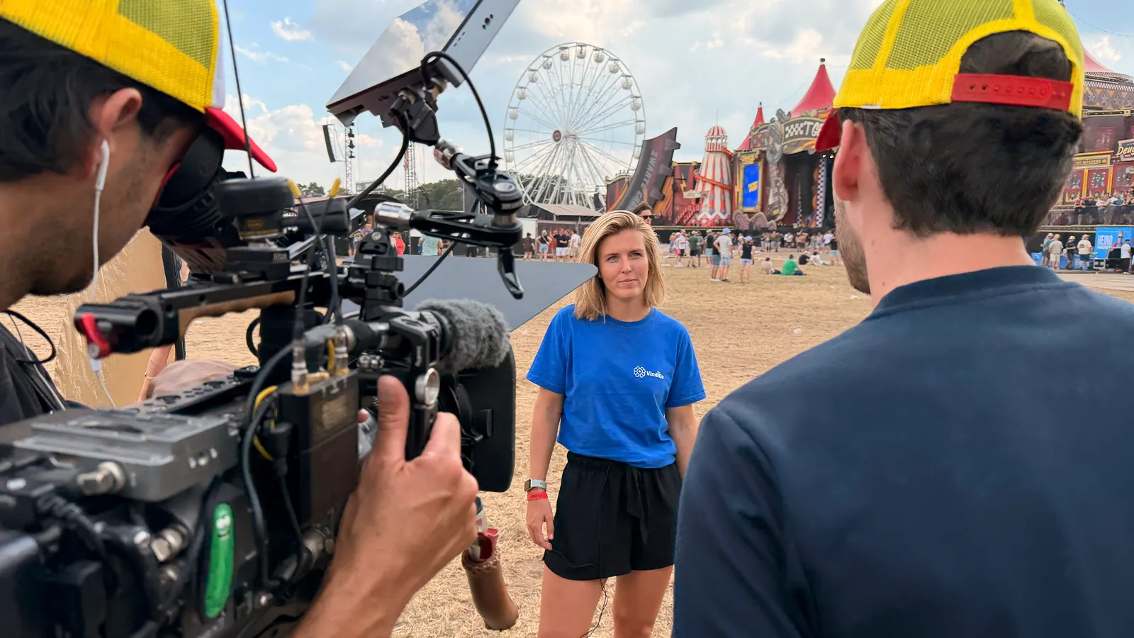 Een vrouw in een blauw shirt wordt gefilmd door een cameraploeg op een buitenfestival, met op de achtergrond een reuzenrad en kleurrijke tenten.
