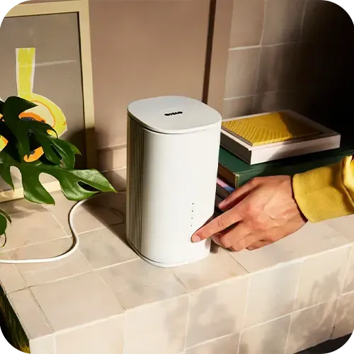 A person in a yellow sleeve presses a button on a sleek, white electronic device on a tiled surface next to a plant and framed artwork.