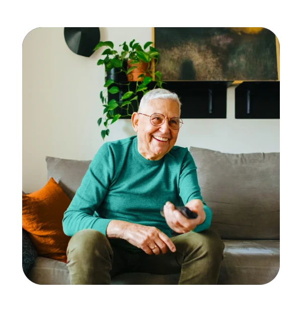 Lachende oudere man in een groene trui die een afstandsbediening voor de tv vasthoudt, zittend op een grijze bank met kussens in een gezellige woonkamer.