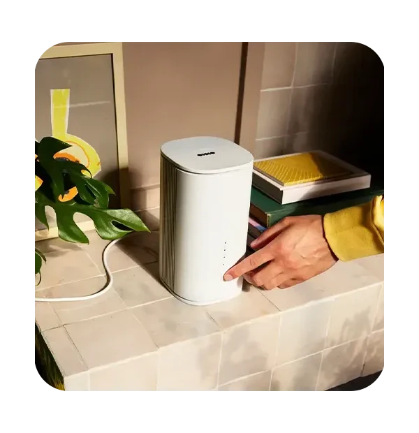 A person in a yellow sleeve presses a button on a sleek, white electronic device on a tiled surface next to a plant and framed artwork.