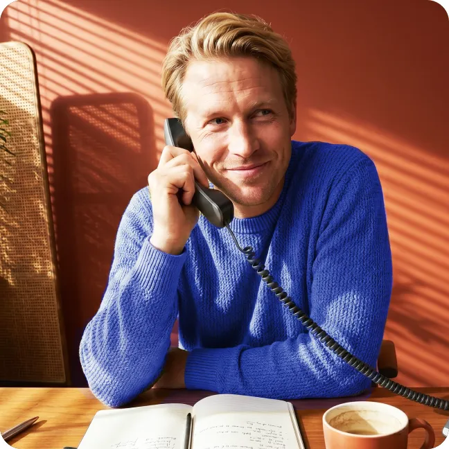 Glimlachende man in een blauwe trui die aan het telefoneren is, met een notitieboek en een koffiekopje op tafel, tegen een warme, zonnige achtergrond.