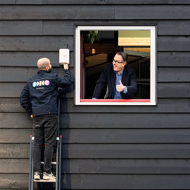 Man op een ladder die een apparaat op een zwarte muur afstelt, terwijl een andere man binnen door een open raam een duim omhoog geeft.