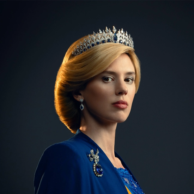 Vrouw met blond haar, een met juwelen bezette tiara, een blauwe jurk en een sierlijke broche, die ernstig kijkt tegen een donkere achtergrond.
