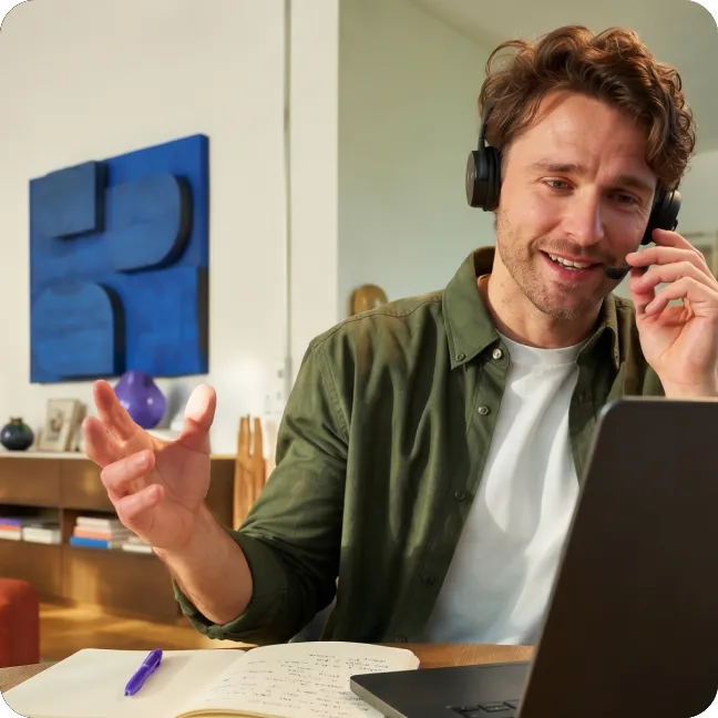 Een man met een koptelefoon spreekt tijdens een videogesprek aan een bureau met een open laptop en pen, met blauwe wandkunst op de achtergrond.