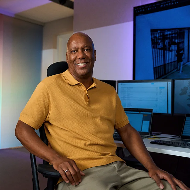 Een lachende man in een geel poloshirt maakt gebaren met zijn handen in een felverlichte kamer met een wazig scherm op de achtergrond.