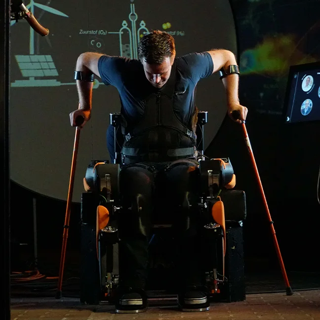 A man in an exoskeleton stands with the help of crutches in front of a projector displaying scientific illustrations.