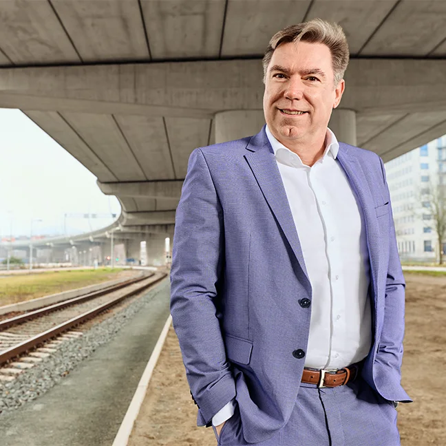 Man in een lichtblauw pak staat zelfverzekerd onder een grote betonnen brug naast spoorrails, met op de achtergrond gebouwen zichtbaar.