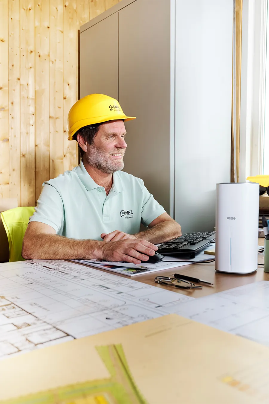 Glimlachende man met een gele veiligheidshelm werkt aan een bureau met bouwtekeningen, een computer en een router in een met hout bekleed kantoor.